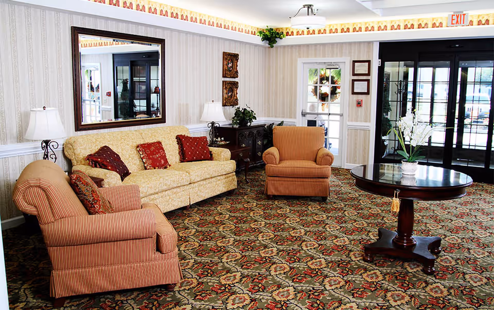 A cozy living room area with a patterned carpet, a yellow sofa with red and patterned cushions, two striped armchairs, a round wooden table with a white orchid plant, two table lamps, a large mirror on the wall, and glass doors leading outside.