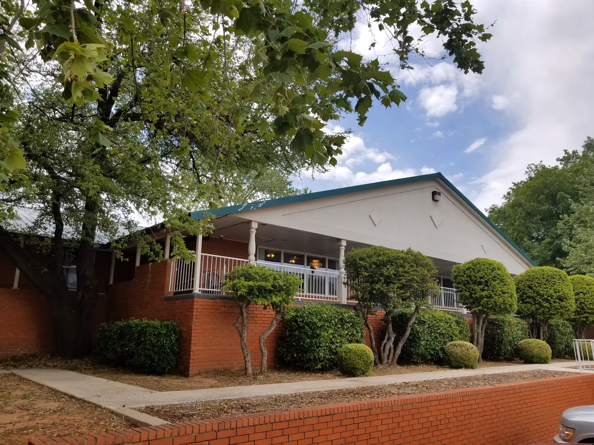 Exterior view of a single-story building with a white triangular roof and red brick walls, surrounded by neatly trimmed bushes and trees under a partly cloudy sky.