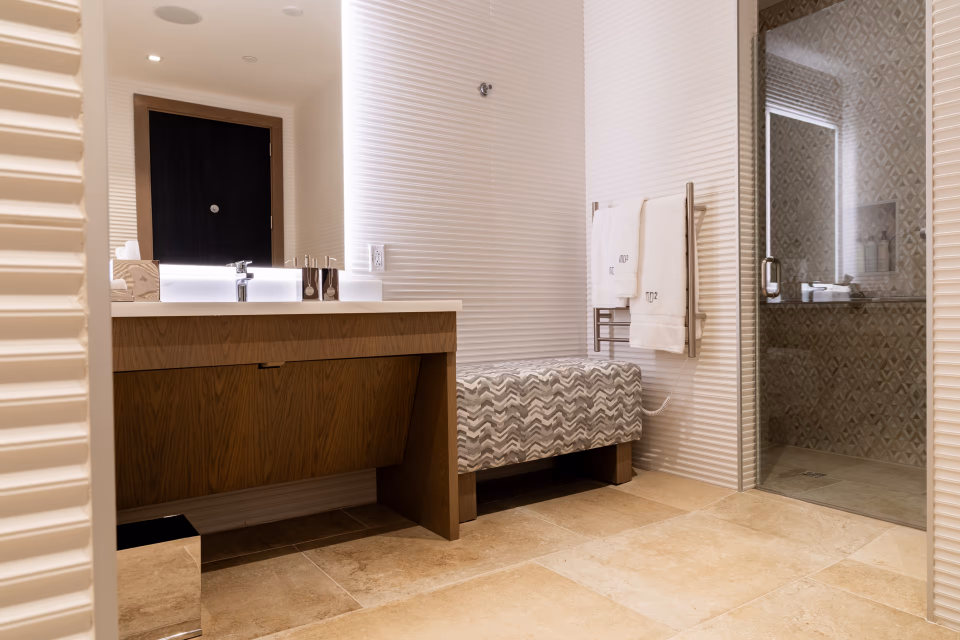 Modern bathroom with a wooden vanity, patterned bench, towel rack, and glass shower.