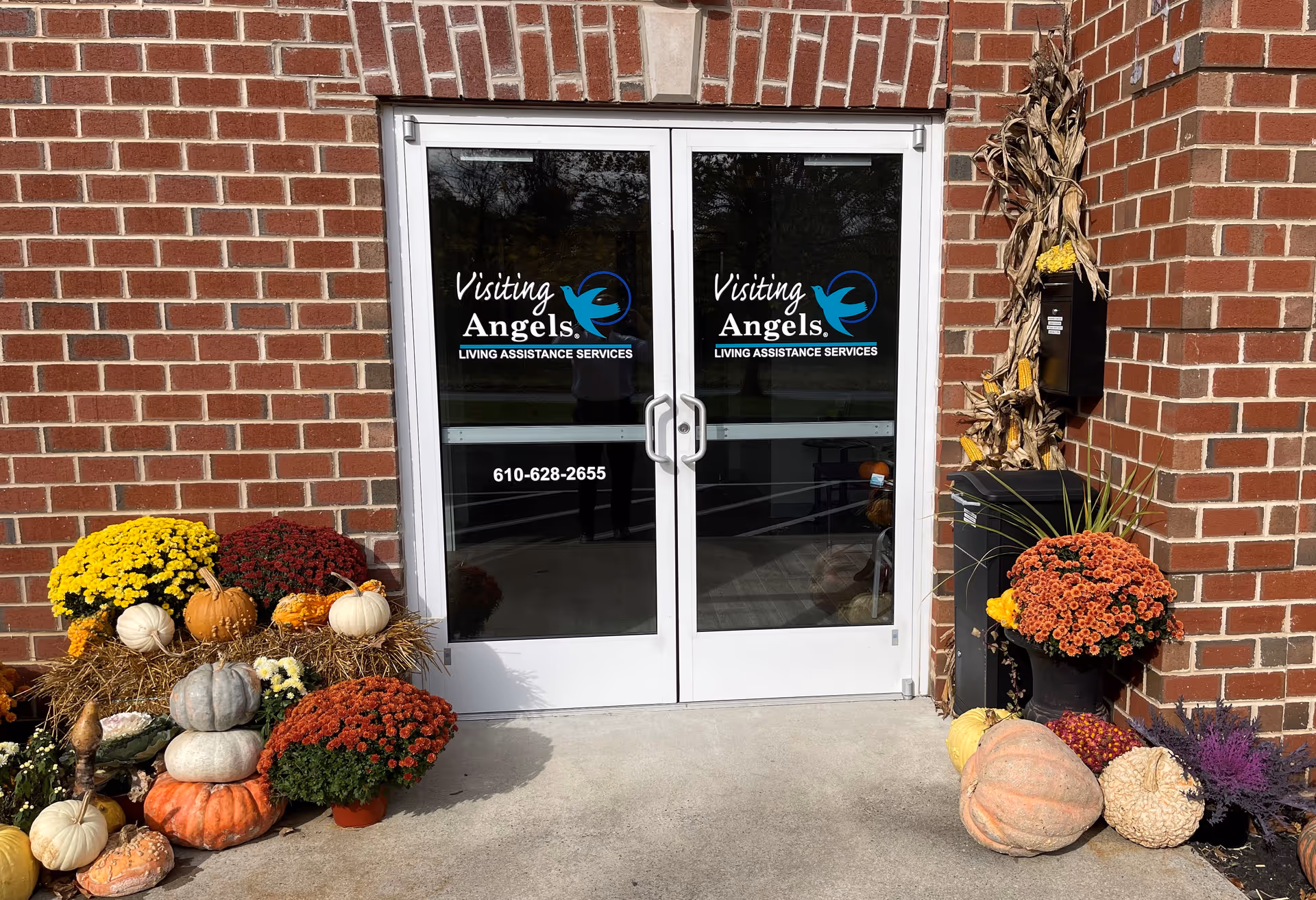 Brick building entrance with glass doors displaying Visiting Angels, autumn pumpkins and mums outside.