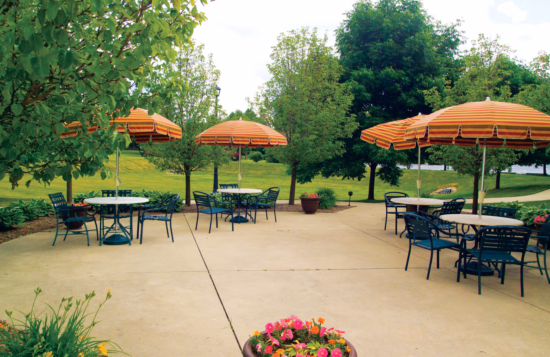 Outdoor patio area with several round tables and chairs, each table shaded by orange and yellow striped umbrellas. The patio is surrounded by green trees, bushes, and a grassy lawn, with a flower pot containing pink and orange flowers in the foreground.
