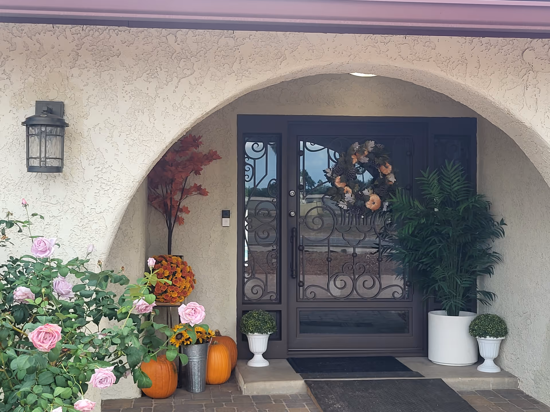 Front entrance of a light-colored building with arched doorway, plants, and pumpkins.