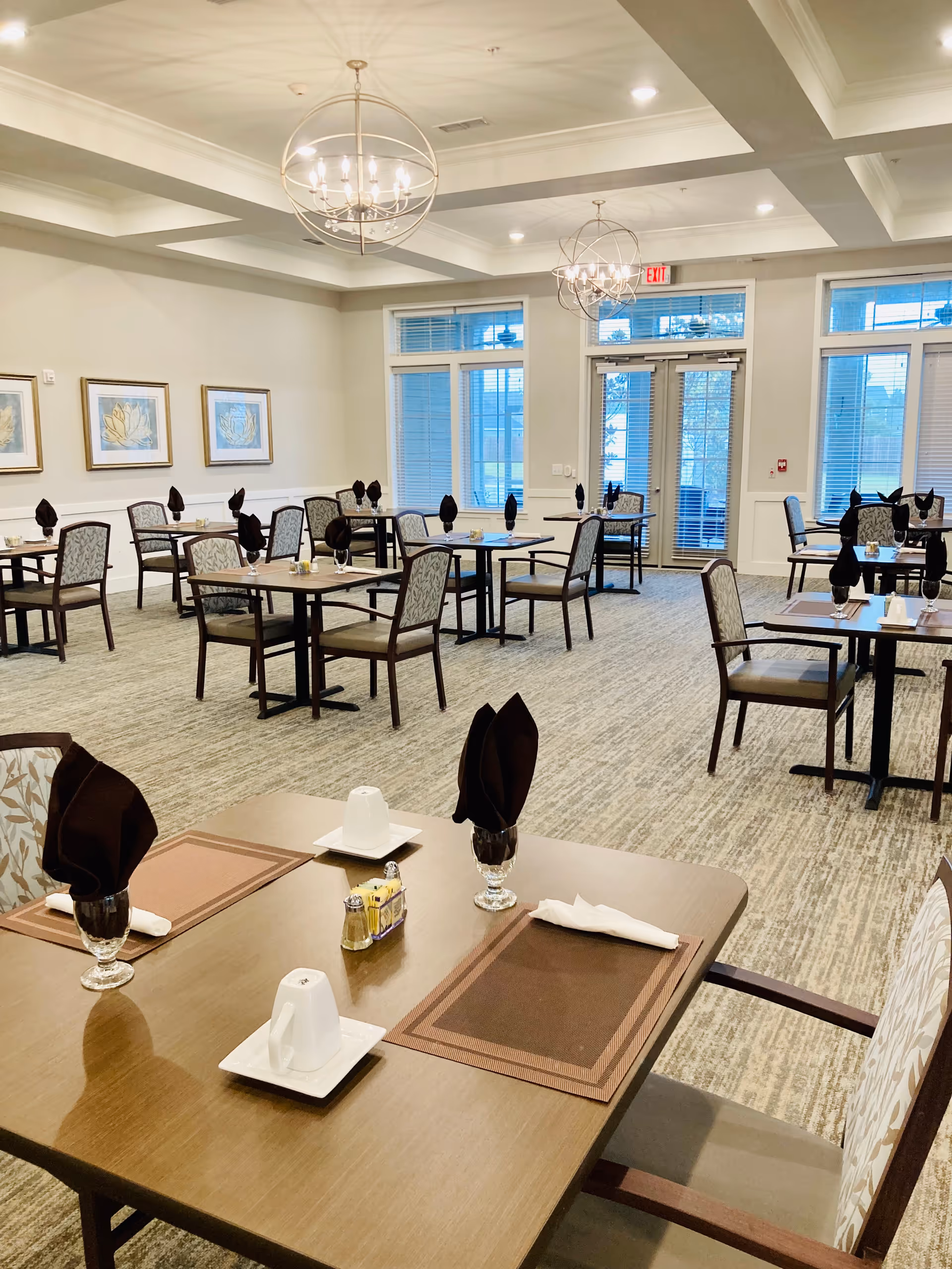 Bright dining room with black-napkin settings, glassware, and arranged tables in a senior living facility.