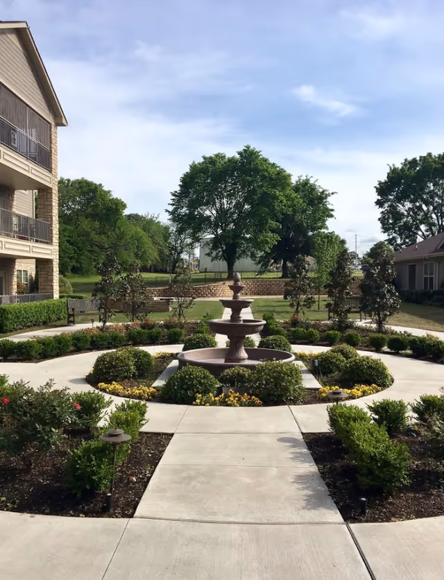 A landscaped outdoor garden area at Legacy Heights Retirement Center featuring a central tiered water fountain surrounded by neatly trimmed bushes and flower beds. Concrete walkways lead to the fountain, with benches and trees in the background under a partly cloudy sky.