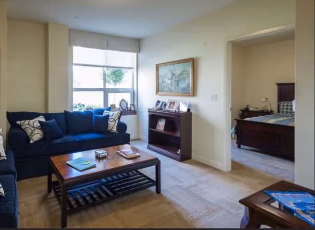 A cozy living room with a dark blue sofa, a wooden coffee table with books and a small decorative box, a window with a white blind, and a small wooden bookshelf displaying framed photos and a painting above it. An adjacent bedroom with a bed and nightstand is visible through an open doorway.