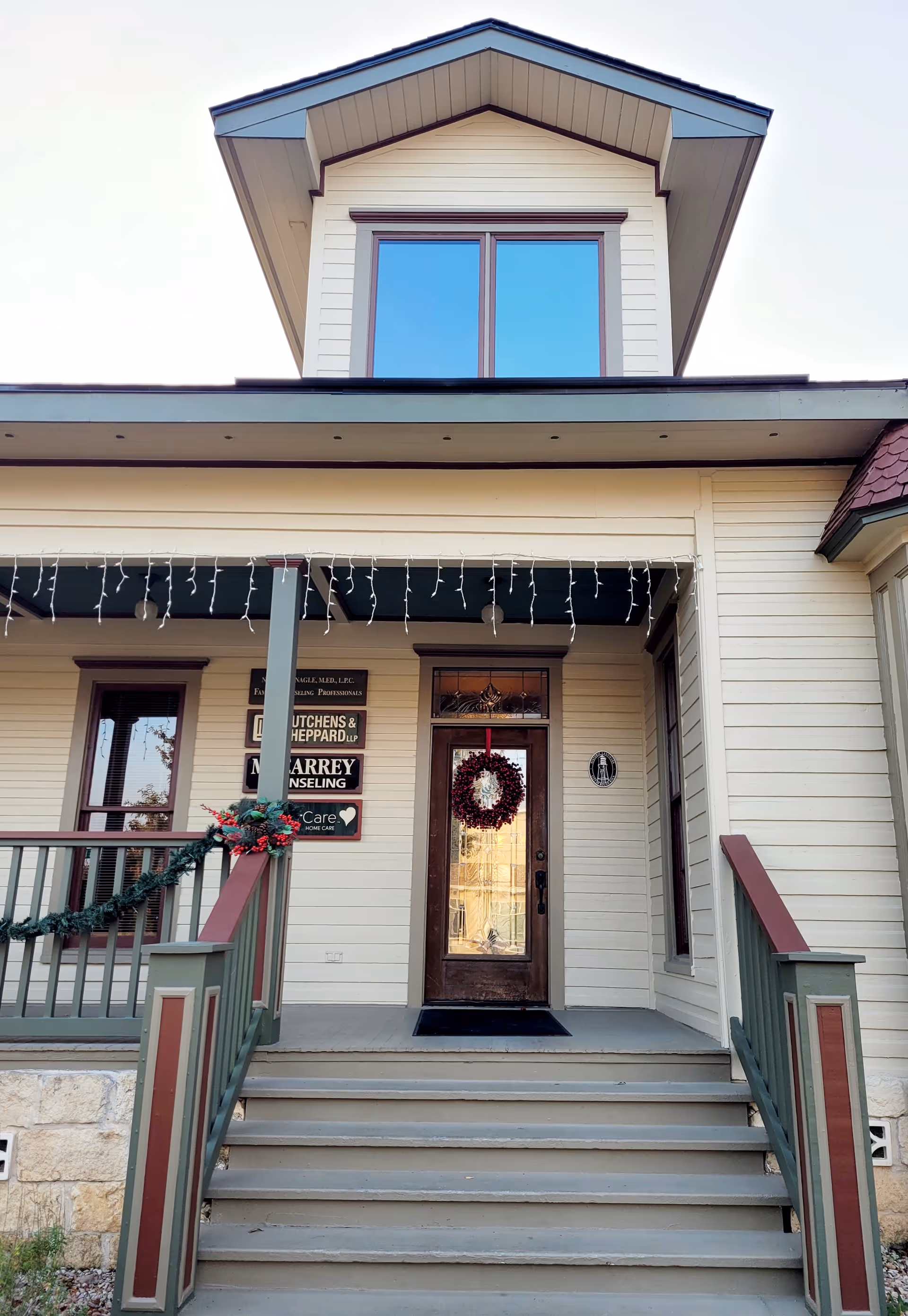 Front entrance of a beige care facility with wreath, stairs, and string lights.