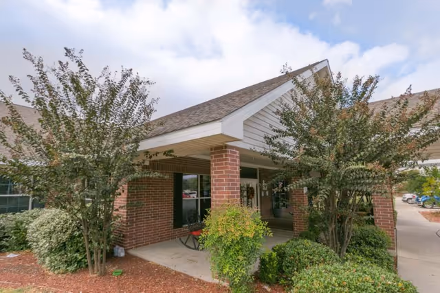 Entrance of a healthcare and rehabilitation facility with a covered porch supported by brick columns, surrounded by green bushes and trees under a partly cloudy sky.