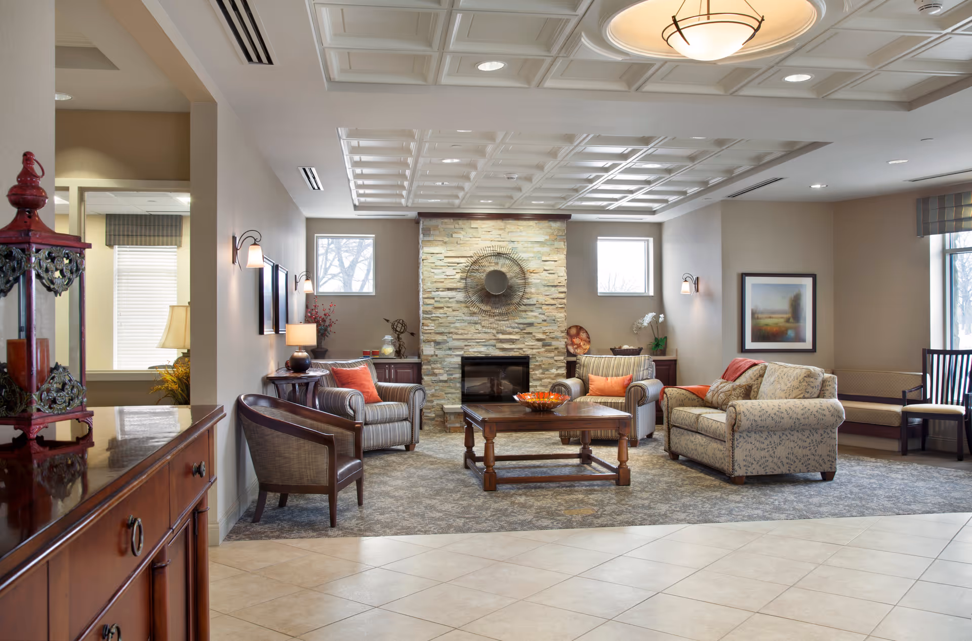 A cozy living room area with a stone fireplace centered between two small windows. The room features a wooden coffee table, two striped armchairs with orange cushions, a floral-patterned sofa with a red throw, and additional seating along the walls. The ceiling has a decorative grid pattern with recessed lighting and a central light fixture. The floor transitions from tiled to carpeted near the seating area. A wooden cabinet with a decorative lantern is visible in the foreground.