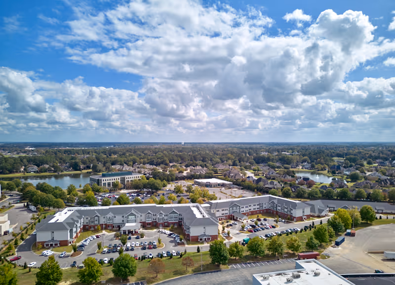 Aerial view of The Crossings at Eastchase facility showing a large, multi-wing building with a parking lot in front, surrounded by trees and residential neighborhoods under a partly cloudy blue sky.