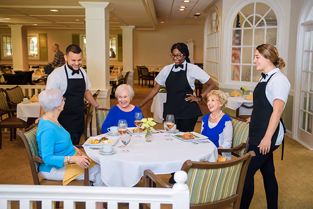 Three elderly women seated around a dining table in a well-lit dining room, smiling and interacting with three waitstaff dressed in white shirts and black aprons. The table is set with plates of food, glasses of water and wine, and a small flower vase. The background shows more dining tables and chairs in a spacious, elegant room.
