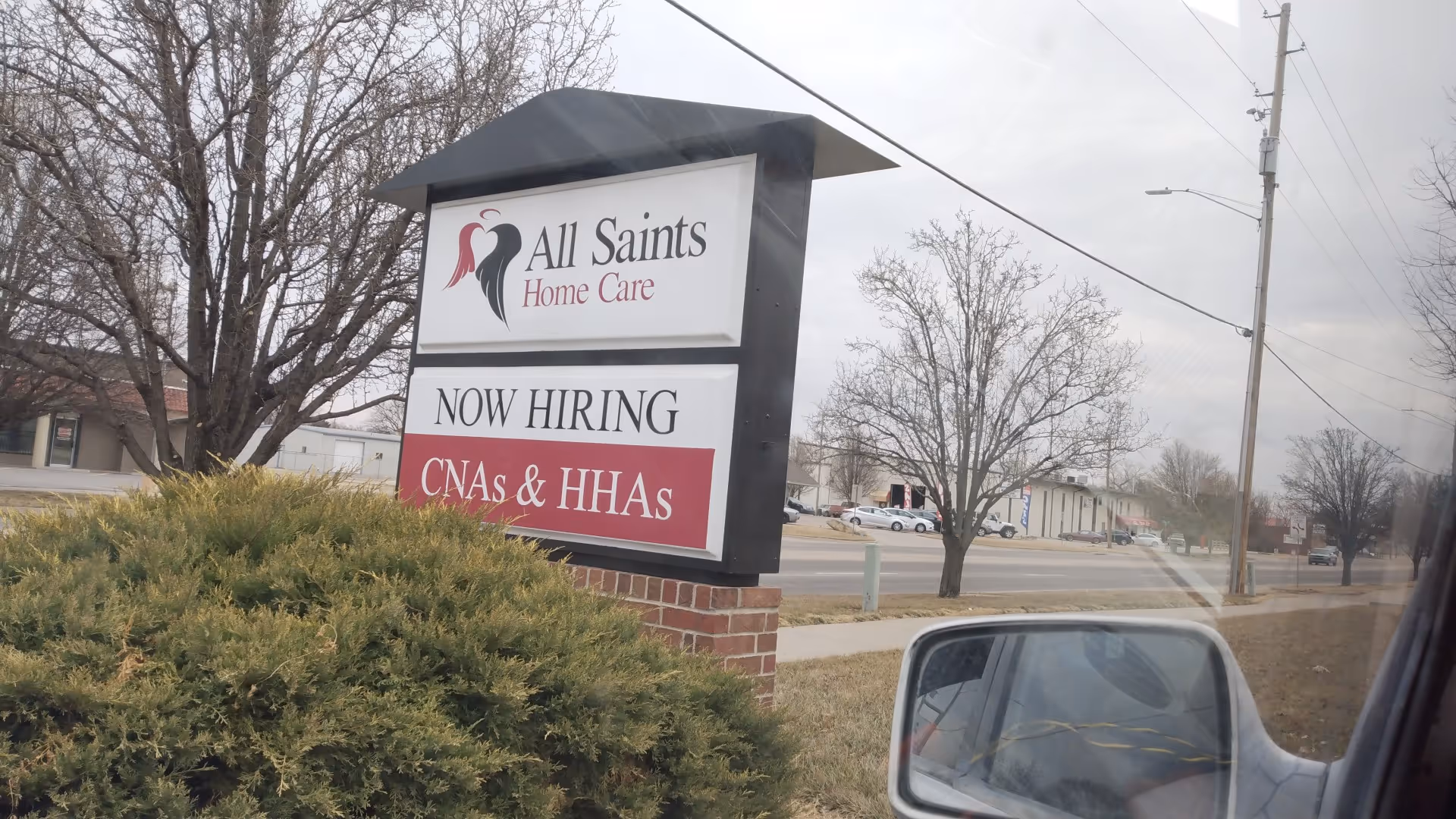 Exterior view of All Saints Home Care sign with 'Now Hiring' banner along the street.