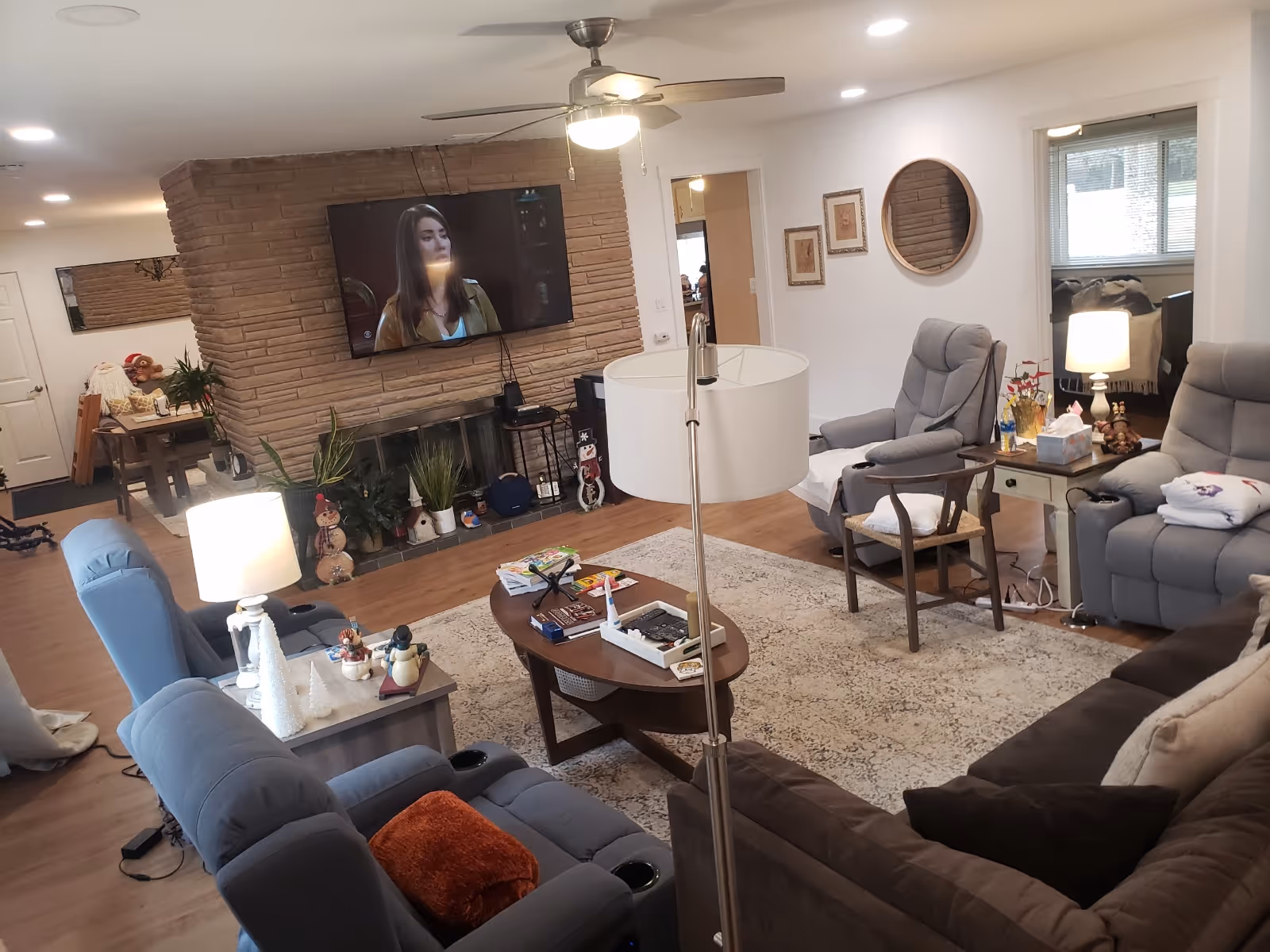 Cozy senior living room with recliners, sofa, coffee table, fireplace, plants, and wall-mounted TV.