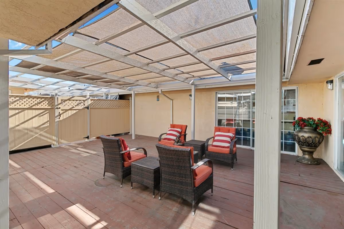 Outdoor patio area with a wooden deck, covered by a pergola with a mesh shade. The patio has four wicker chairs with red cushions and striped pillows arranged around two small matching tables. There is a large decorative planter with red flowers near the sliding glass doors leading inside the building.