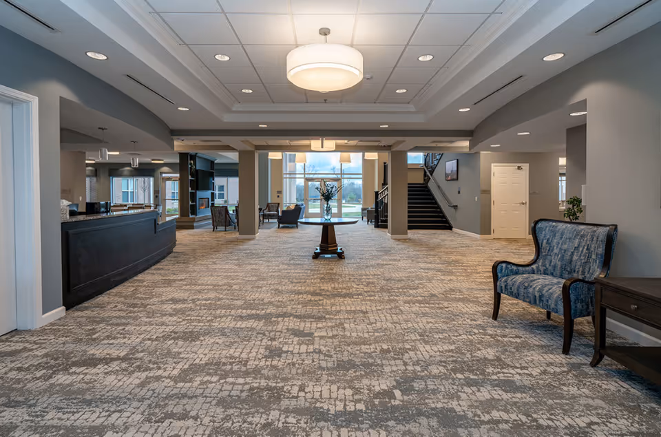 Spacious senior living lobby with a central round table, seating areas, and a staircase.