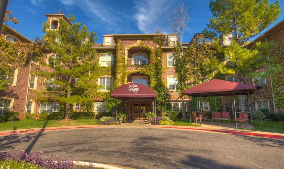 Brick assisted-living building with ivy, trees, and outdoor seating beneath red canopies.
