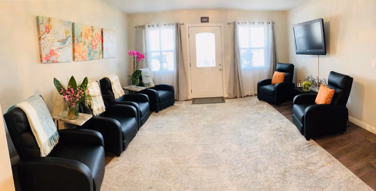 Well-lit common lounge with black recliner chairs along both walls, a wall-mounted TV, artwork, and a central light-colored rug facing an entry door.