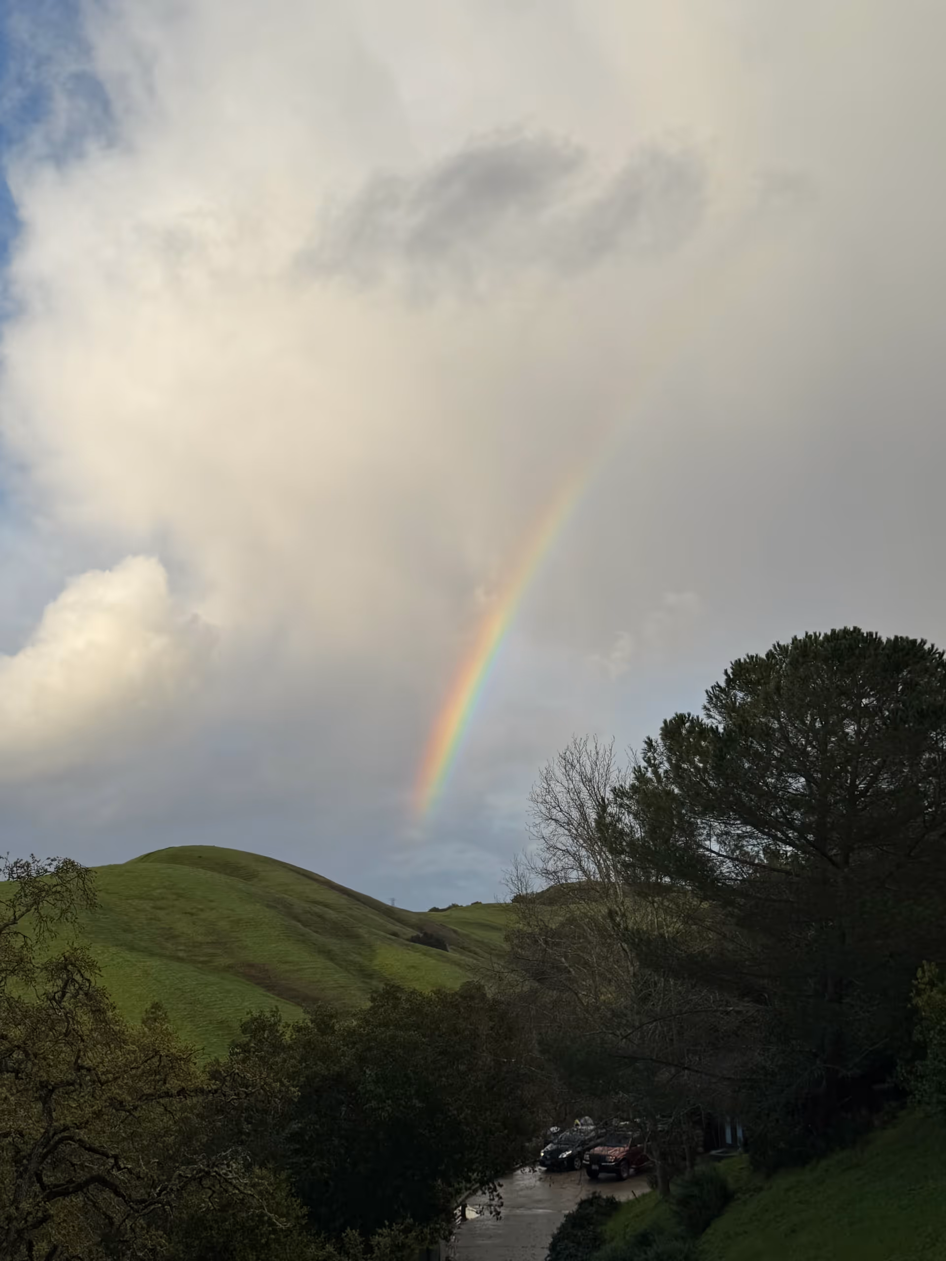 Rainbow over green rolling hills and trees, with a parking lot below.