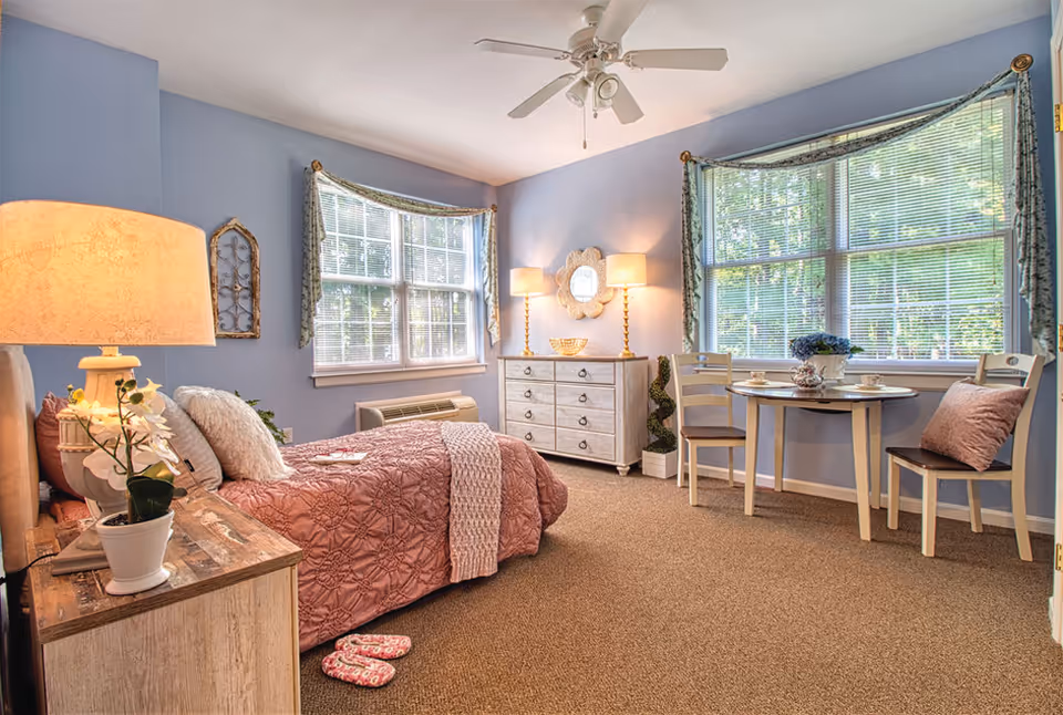 Blue-carpeted bedroom with pink quilted bed, dresser, lamps, and a small dining table.