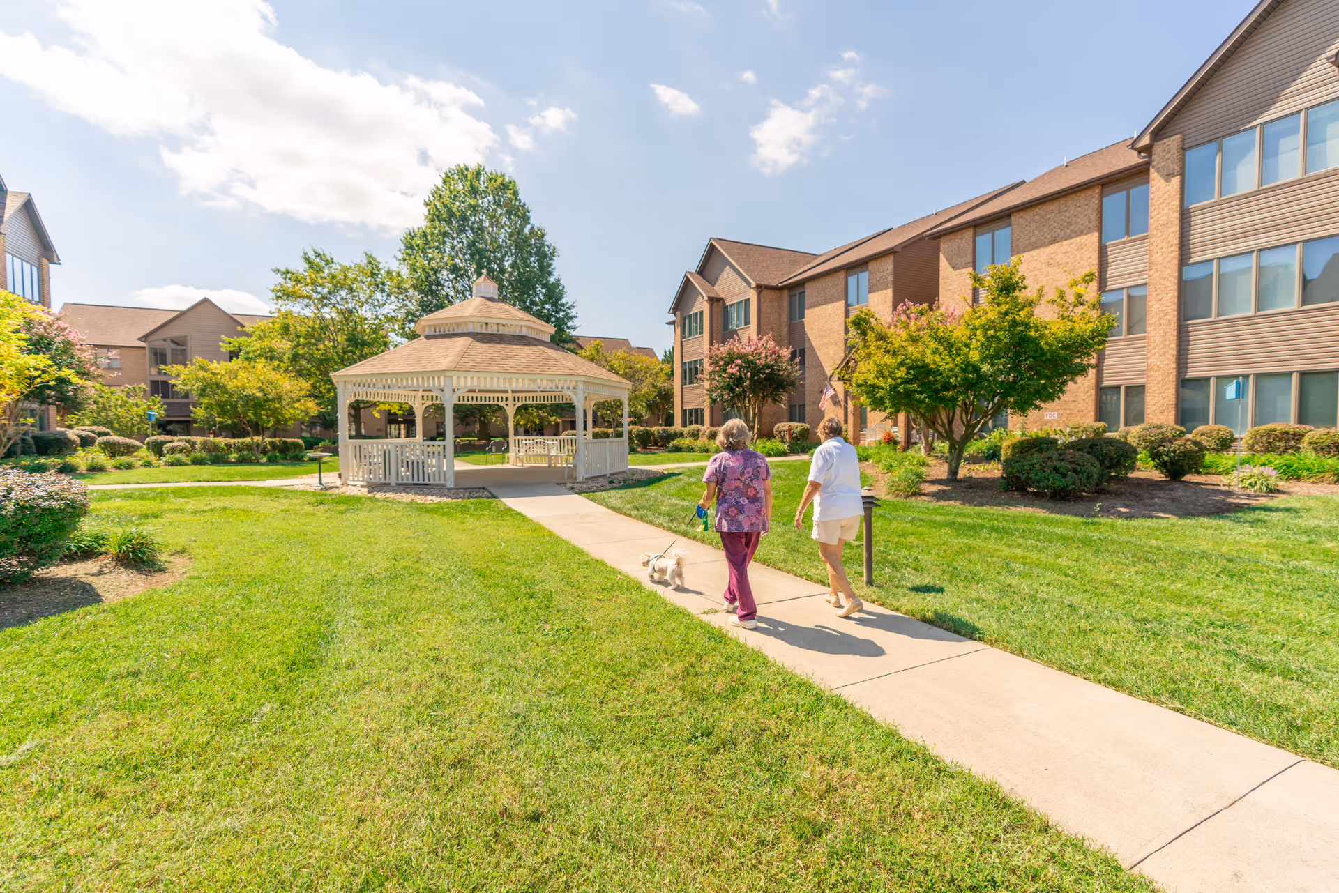 Two residents walk a small dog along a sunny courtyard path past a gazebo.