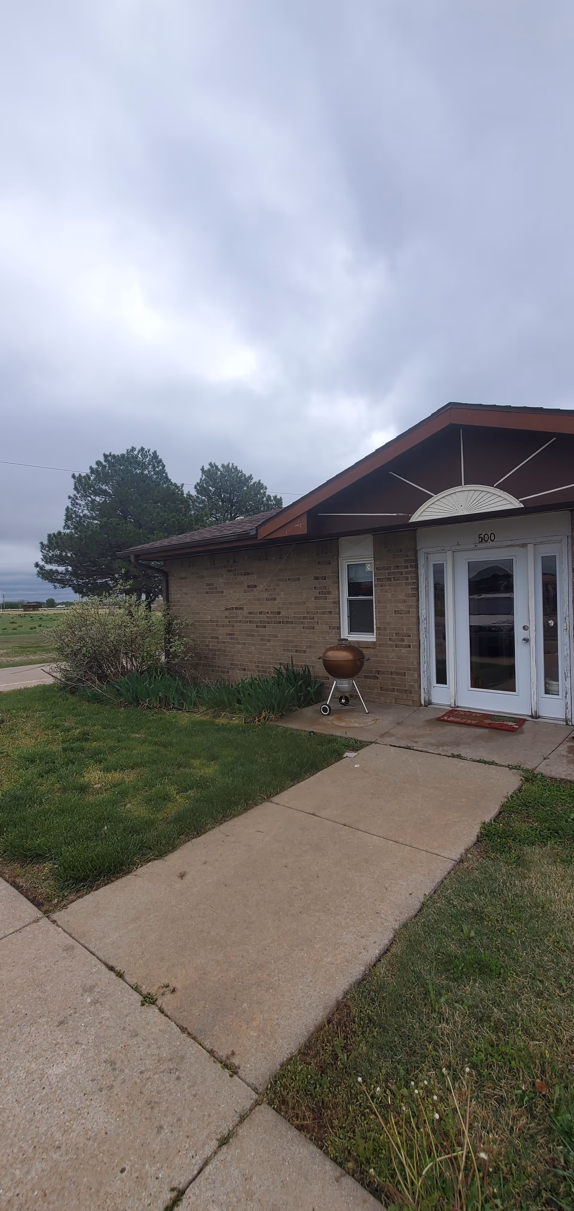 Brick senior living facility entrance with glass doors, sidewalk, and a barbecue grill outside.