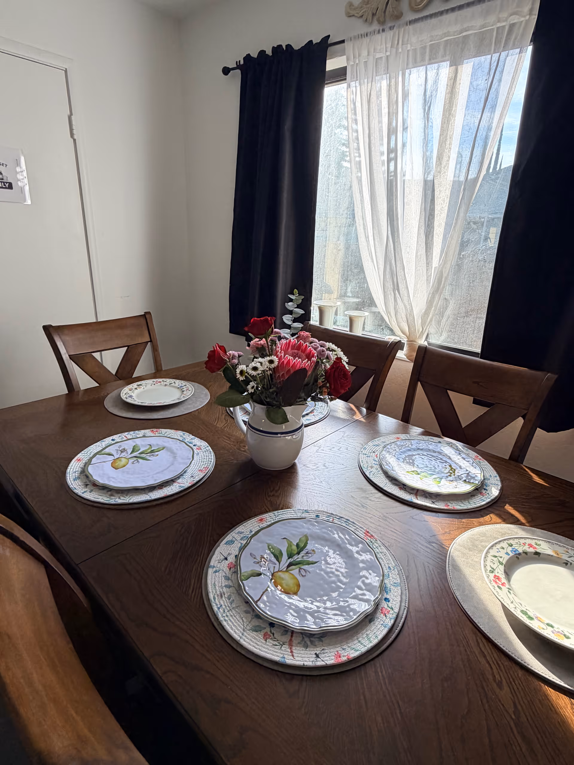 Wooden dining table set with plates and a floral centerpiece by window.