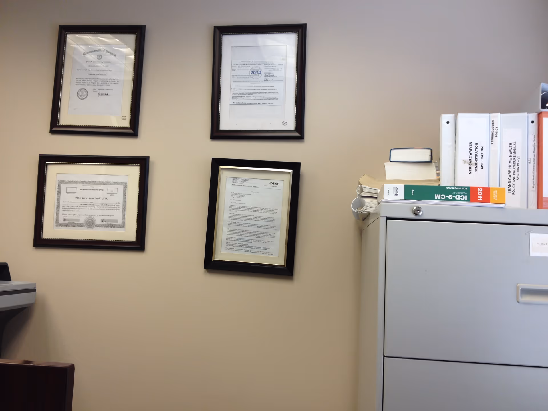 Office corner with framed certificates on the wall and a filing cabinet.