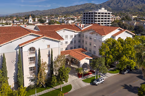 Aerial view of a multi-story senior living building with red-tile roofs, a covered entrance, surrounding trees, street and distant mountains.