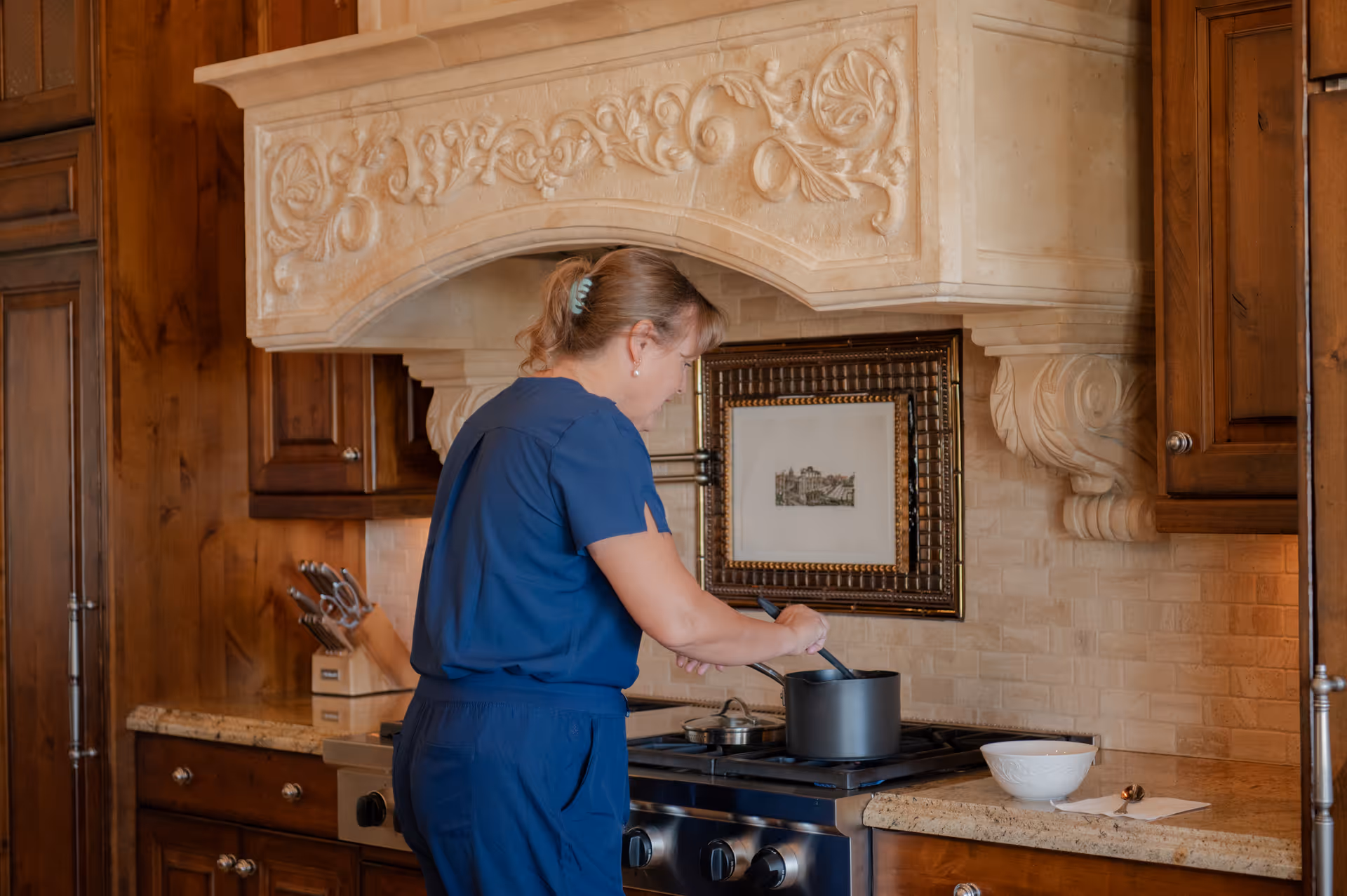 A person in blue scrubs cooks at a stove in an ornate kitchen with wooden cabinets.
