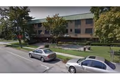 Exterior view of Mayfair Care Center with parked cars, trees, and a green front lawn.
