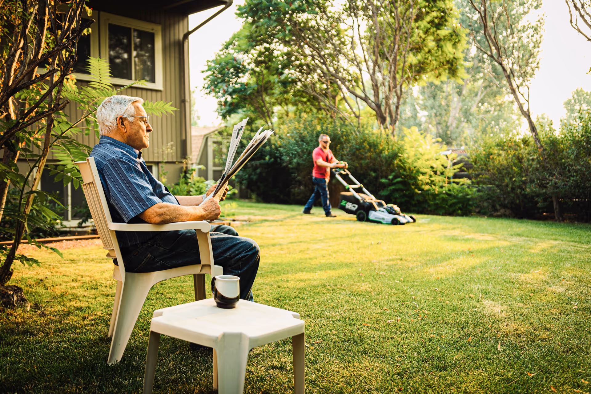Older man sits outdoors on a lawn chair reading a newspaper with a mug nearby.