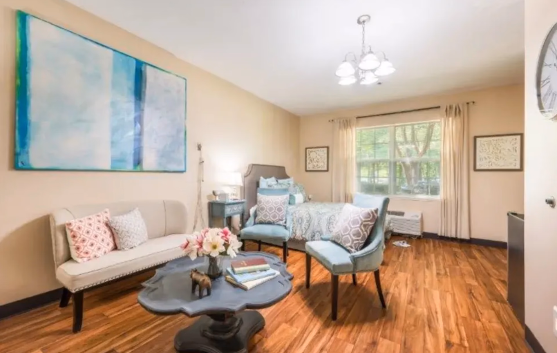 A bright and cozy room with wooden flooring featuring a bed with patterned bedding, two blue cushioned chairs with decorative pillows, a small beige sofa with pillows, a dark wooden coffee table with books and a flower vase, a large window with beige curtains, and light-colored walls adorned with framed artwork and a large abstract painting.
