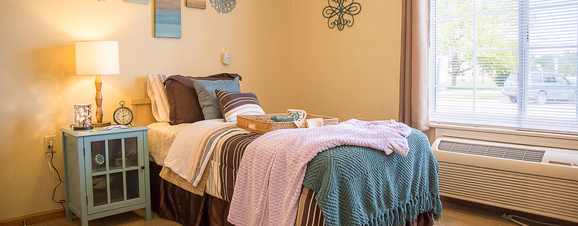 A cozy bedroom featuring a single bed with multiple pillows and layered blankets in shades of brown, teal, and pink. A light blue nightstand with a lamp, alarm clock, and small decorative items sits beside the bed. The room has beige walls adorned with wall art and a large window with blinds letting in natural light. Below the window is a heating and air conditioning unit.