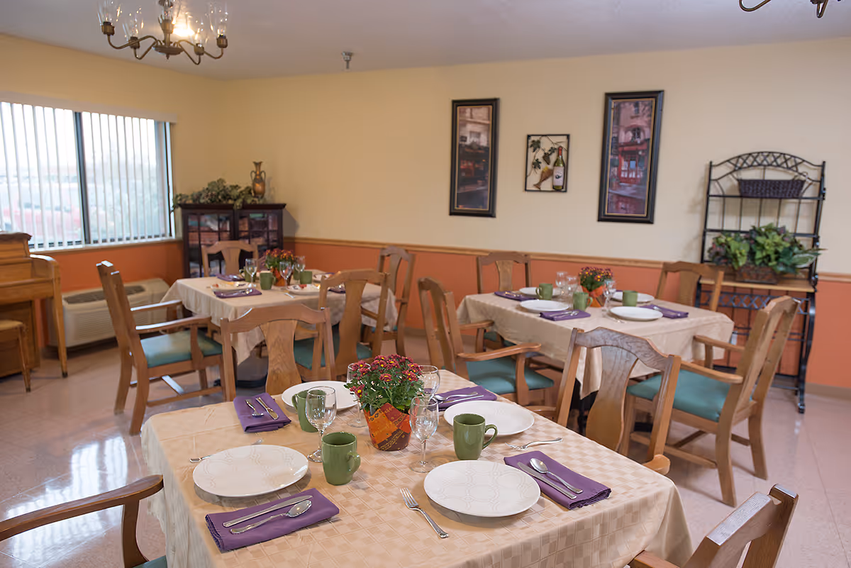 A dining room in a senior living facility with several tables covered in beige tablecloths, each set with white plates, green mugs, silverware wrapped in purple napkins, and small flower centerpieces. Wooden chairs with green cushions surround the tables. The room has light-colored walls with framed artwork and a window with vertical blinds letting in natural light. There is a wooden piano and a black metal shelf with plants in the background.