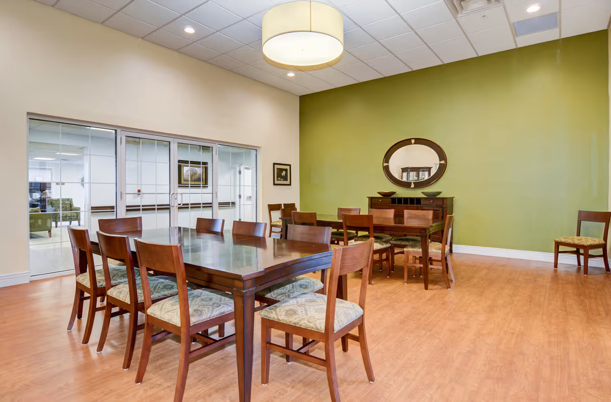 A dining room with two wooden tables surrounded by wooden chairs with patterned cushions. The room has a green accent wall with a round mirror and a wooden sideboard beneath it. The floor is wood, and there is a large circular ceiling light fixture. Glass doors lead to another room with seating visible through the glass.