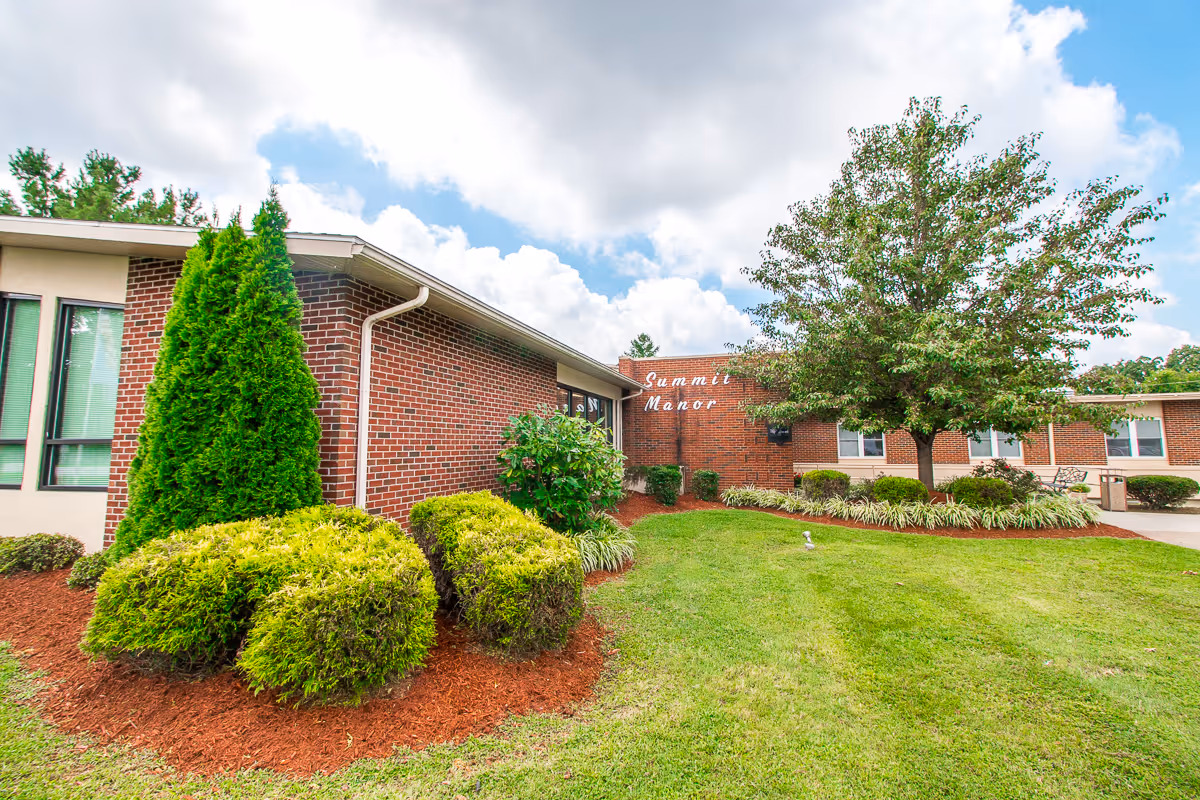 Exterior view of a brick building with well-maintained landscaping including green bushes, a tree, and a lawn under a partly cloudy sky. The building has windows and a sign that reads 'Summit Manor'.