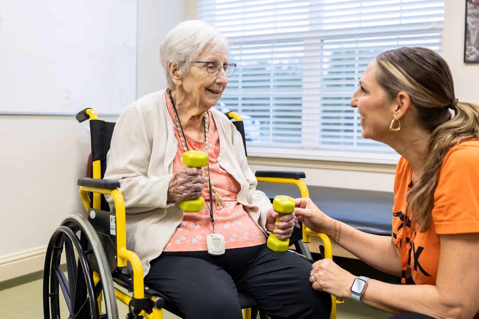 An elderly woman in a wheelchair holding green dumbbells is smiling and exercising with the assistance of a caregiver who is kneeling beside her and encouraging her. They are in a well-lit room with a window and a whiteboard in the background.