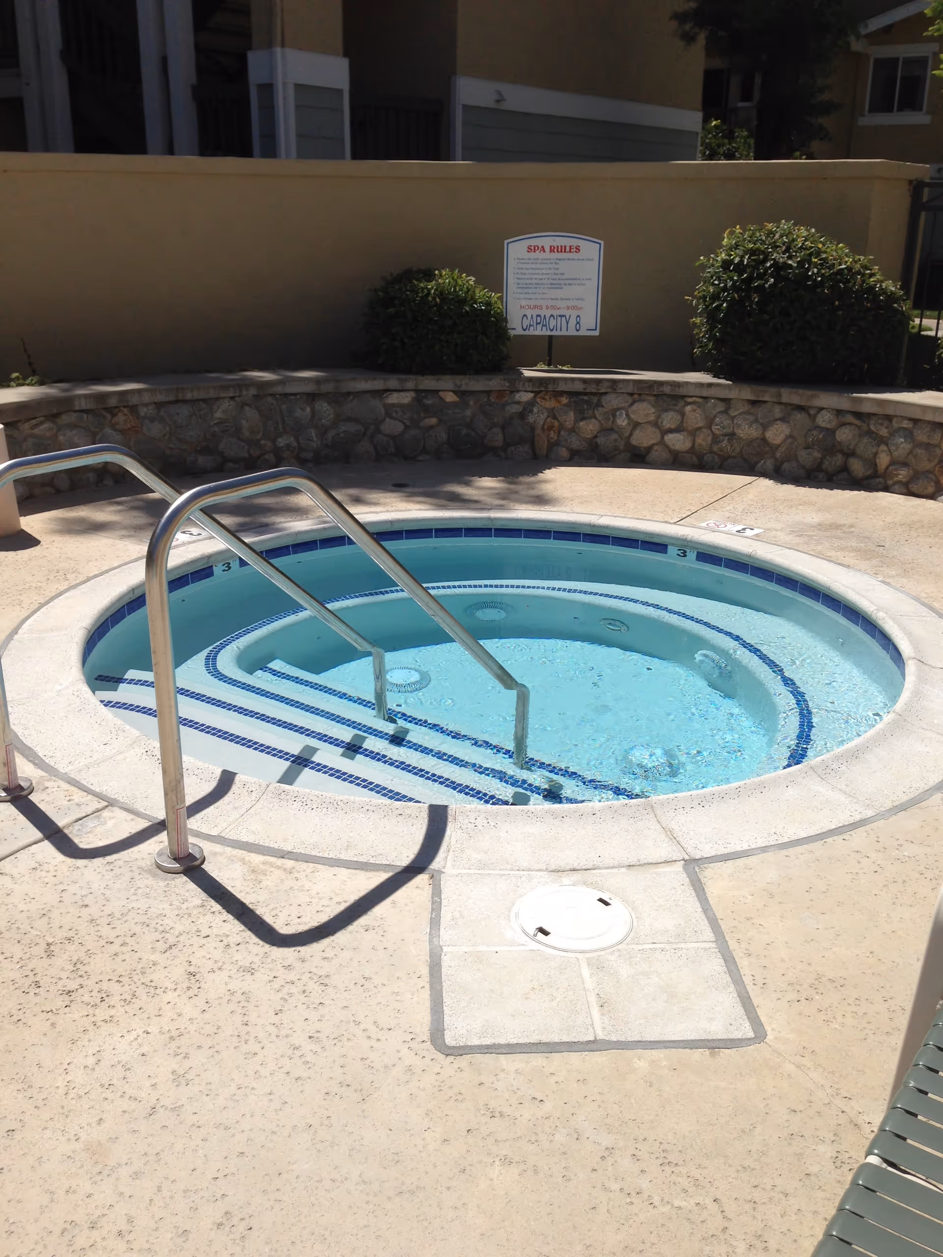 Circular outdoor hot tub with metal handrails and tiled steps in a courtyard beside a stone wall and a spa rules sign.