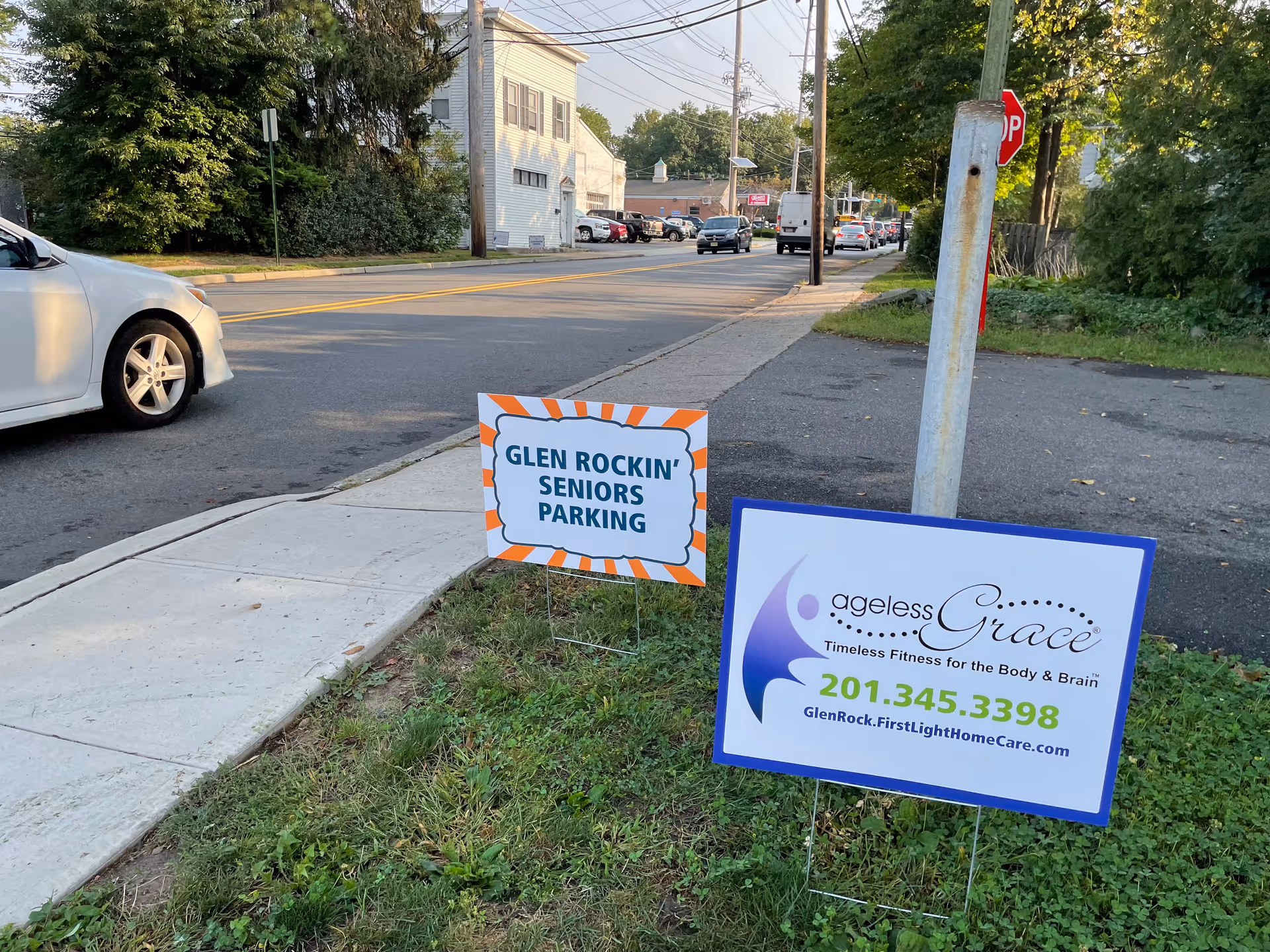 Outdoor street scene with lawn signs advertising Glen Rock FirstLight Home Care and senior parking.