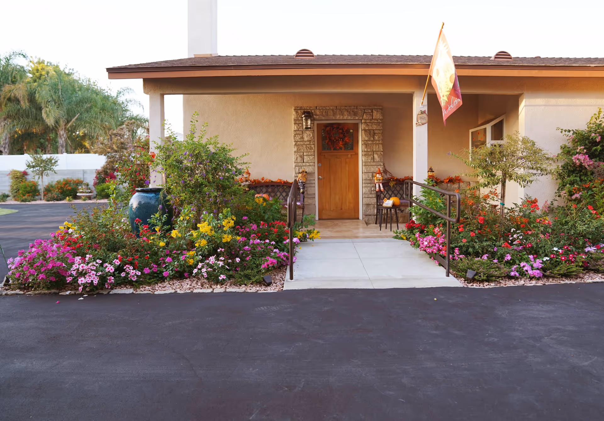Front entrance of a single-story building with a wooden door framed by stone, surrounded by colorful flower beds and greenery. There is a concrete ramp with handrails leading up to the door, and a decorative flag is hanging on the right side of the porch.