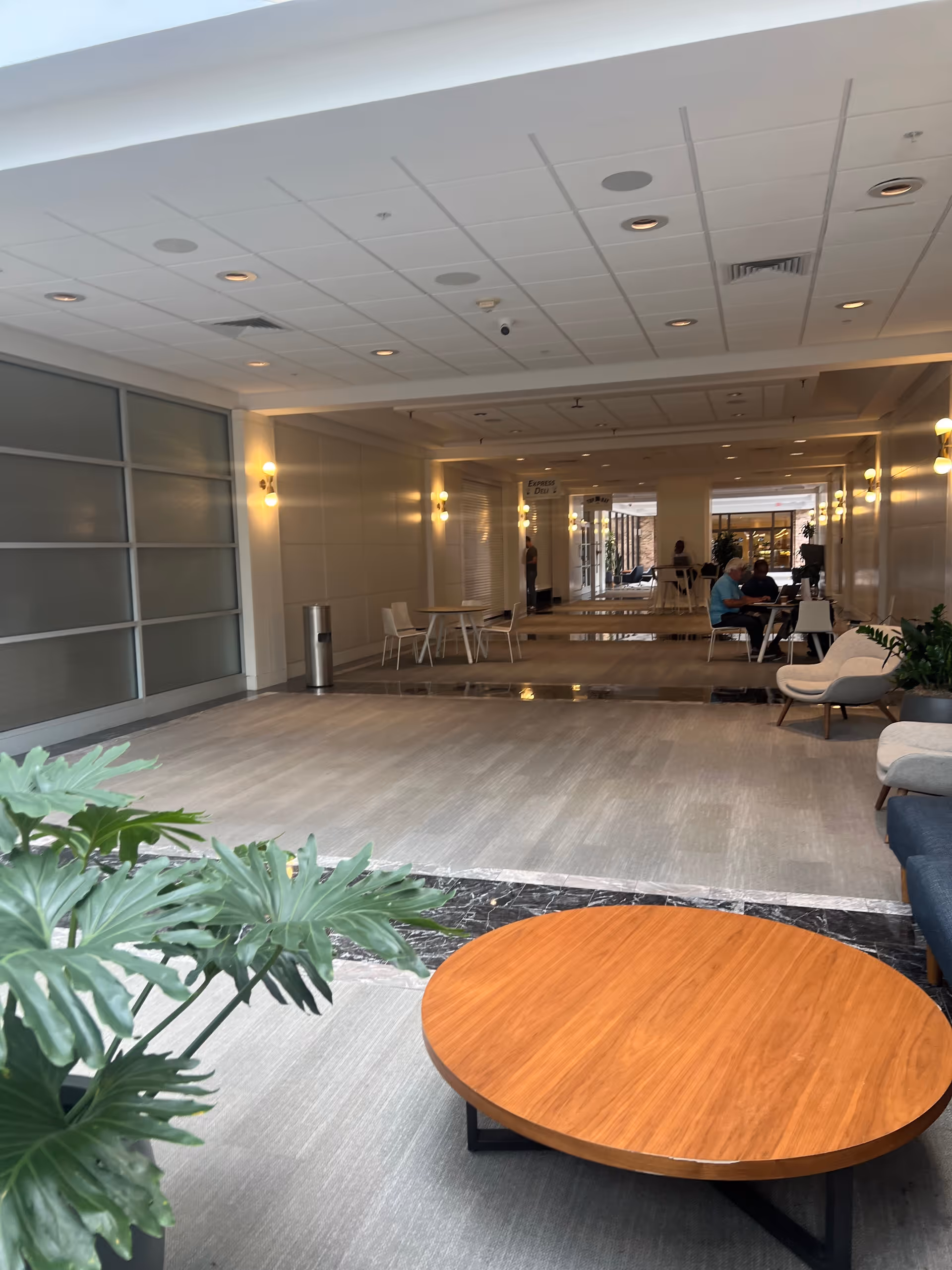 Spacious indoor lounge with round coffee table, plants, and residents seated at distance.