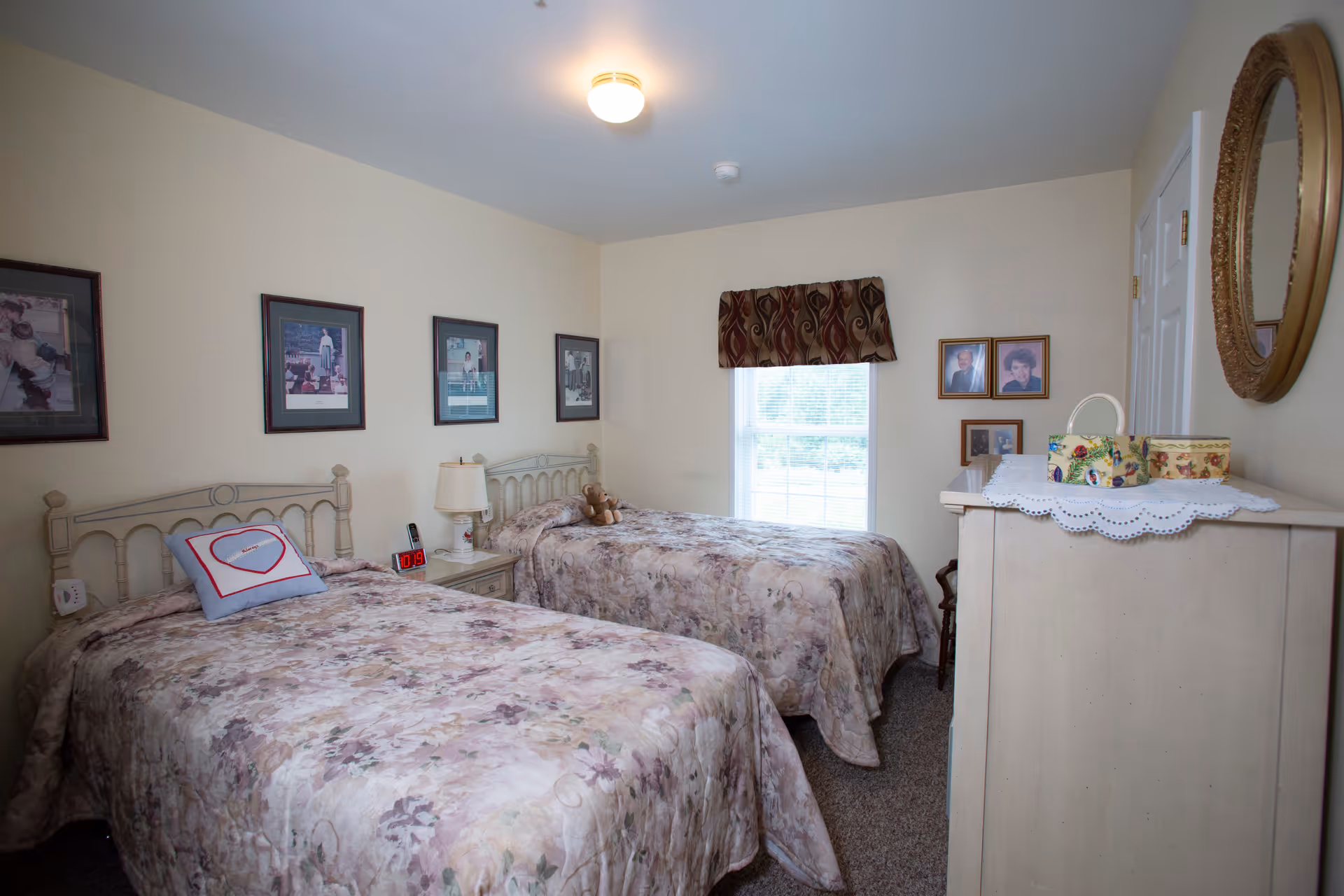 A cozy bedroom with two twin beds covered in floral bedspreads. Between the beds is a nightstand with a lamp and a digital clock. The walls are decorated with framed pictures and a window with a patterned valance lets in natural light. A dresser with decorative boxes and a round mirror is visible on the right side of the room.