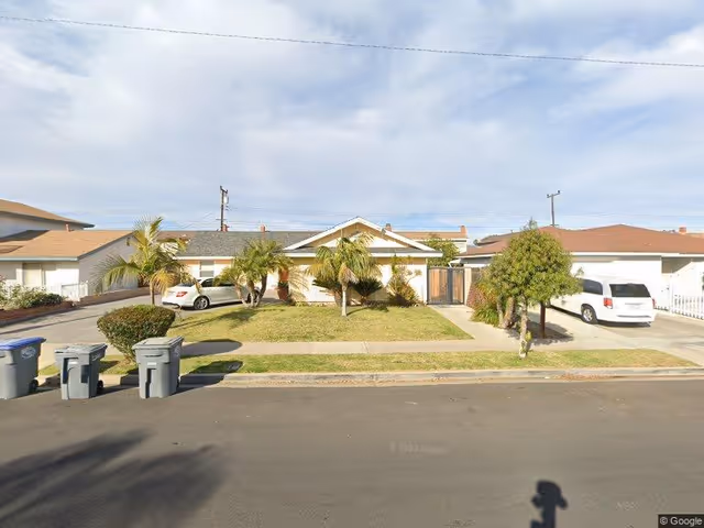 Front exterior of a single-story suburban house with a lawn, palm trees, driveways, parked cars, and trash bins at the curb.