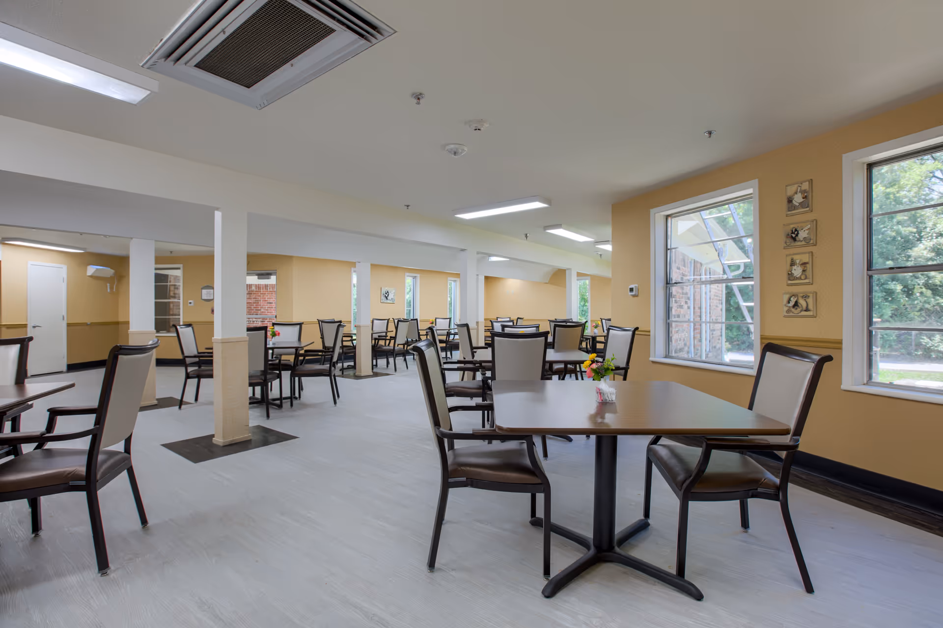 Spacious communal dining room with many tables and chairs, beige walls, and large windows.