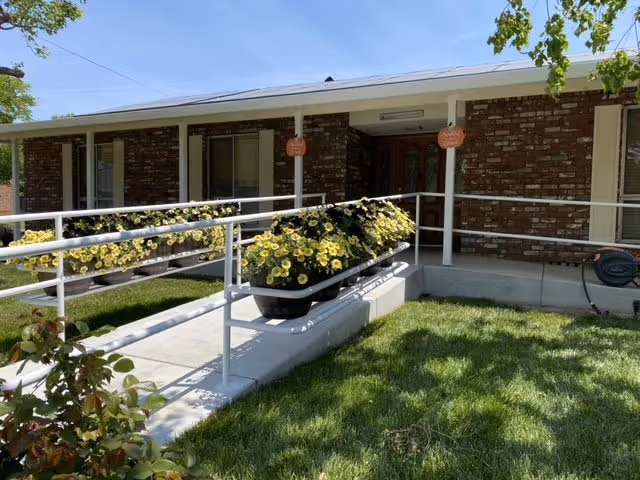 Single-story brick building entrance with a wheelchair ramp, planter boxes of yellow flowers, and a small front lawn.