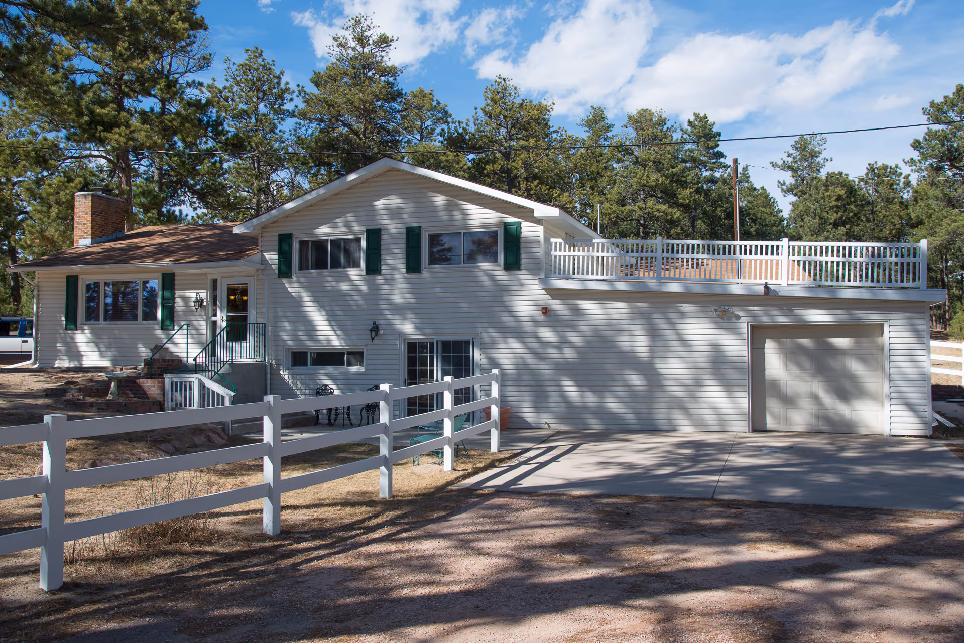 Exterior view of a white two-story house with green shutters, a brick chimney, and a white fence in front. The house is surrounded by tall pine trees under a partly cloudy blue sky.