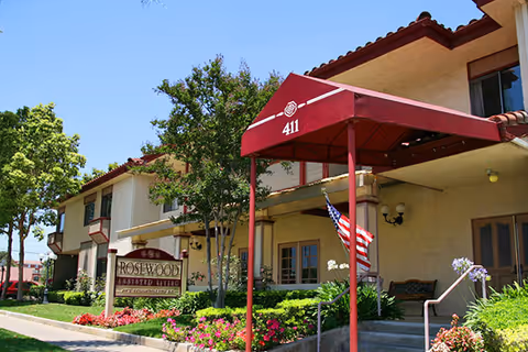 Front entrance of Rosewood Assisted Living — a two-story beige building with a maroon awning marked "411", an American flag, and landscaped flower beds.