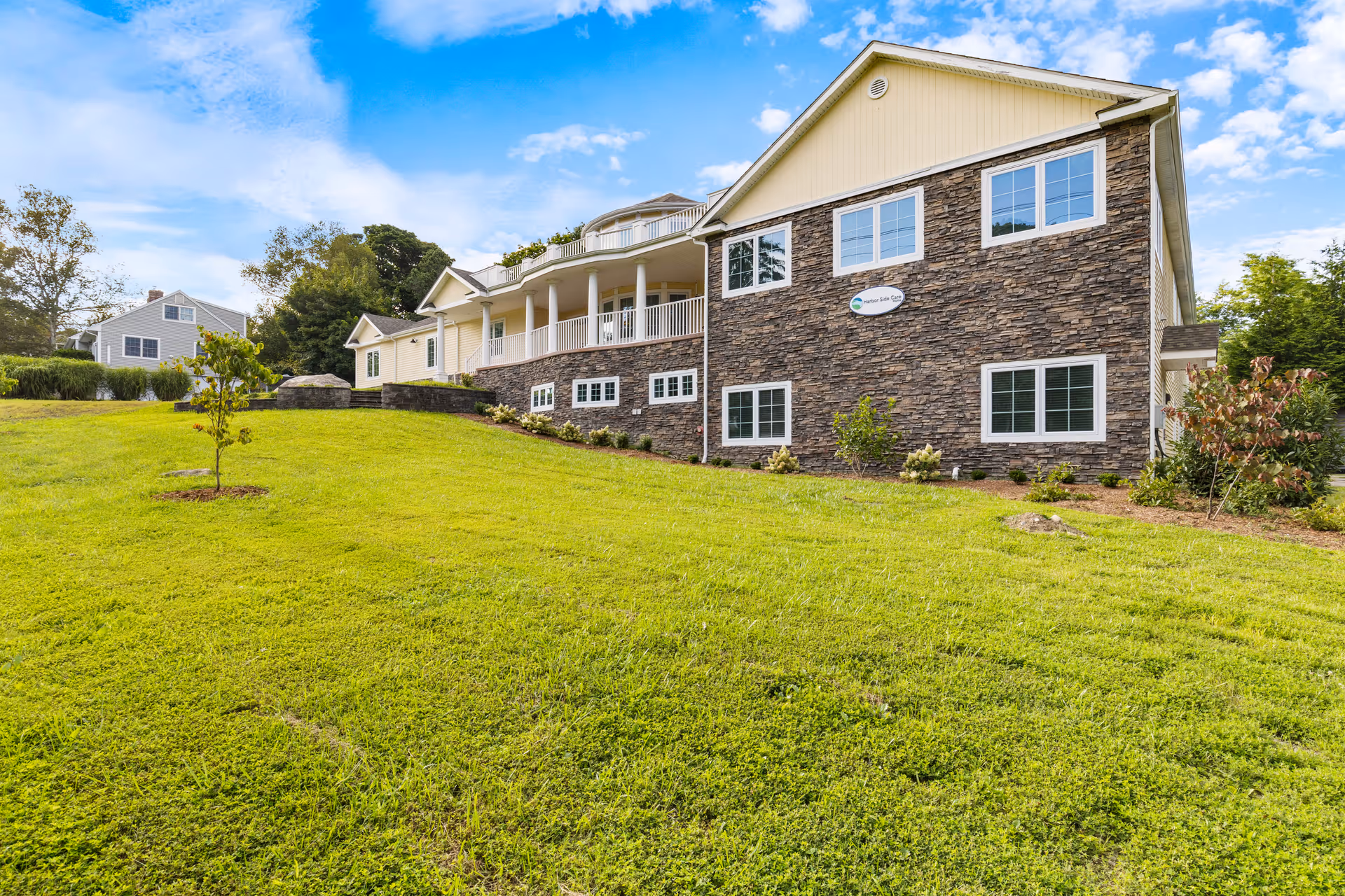 Exterior view of a residential care home building with stone and beige siding, multiple windows, a porch with white railings, and a well-maintained green lawn under a partly cloudy blue sky.