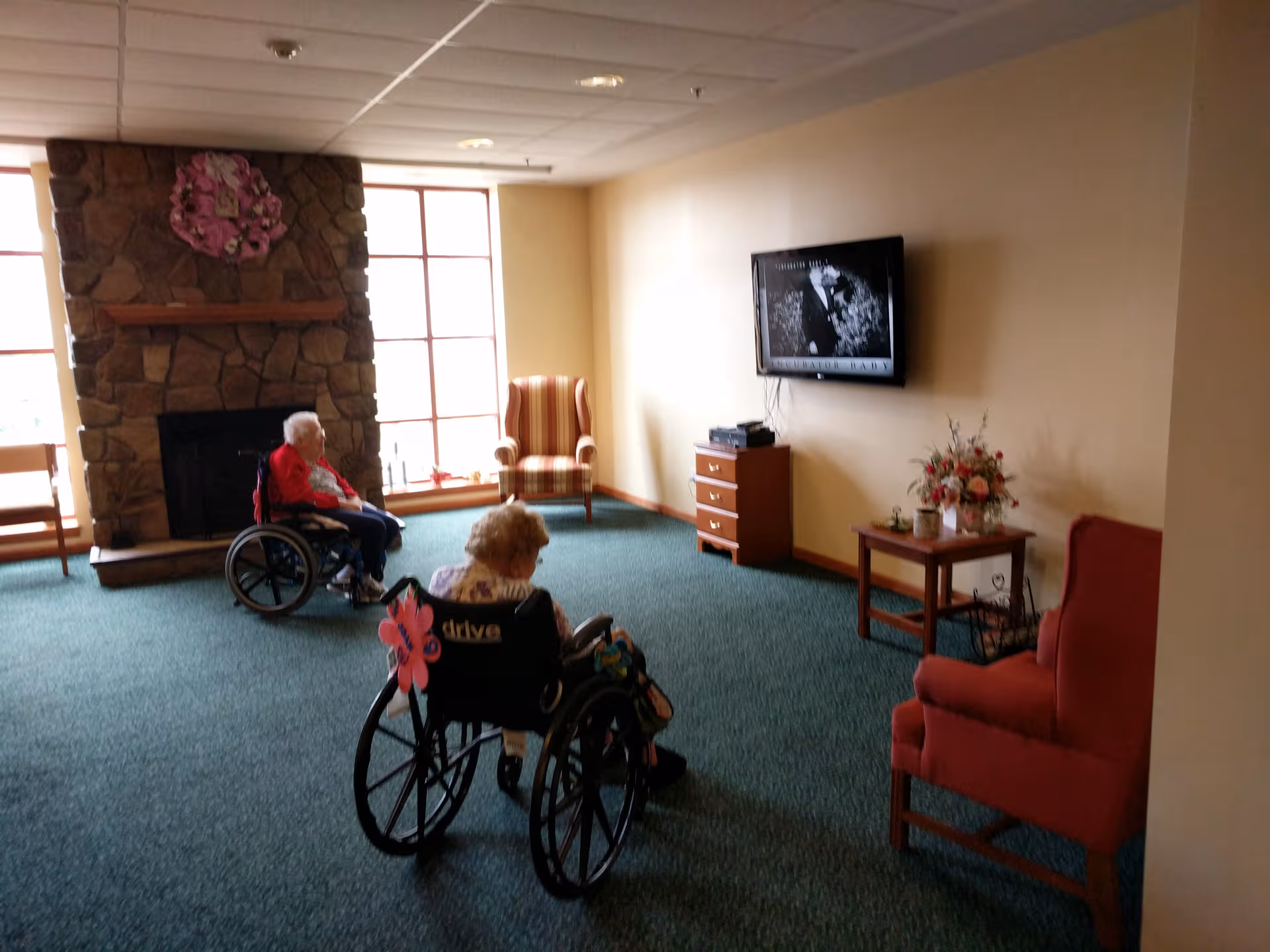 Two elderly residents in wheelchairs watch TV in a cozy living room with a stone fireplace.
