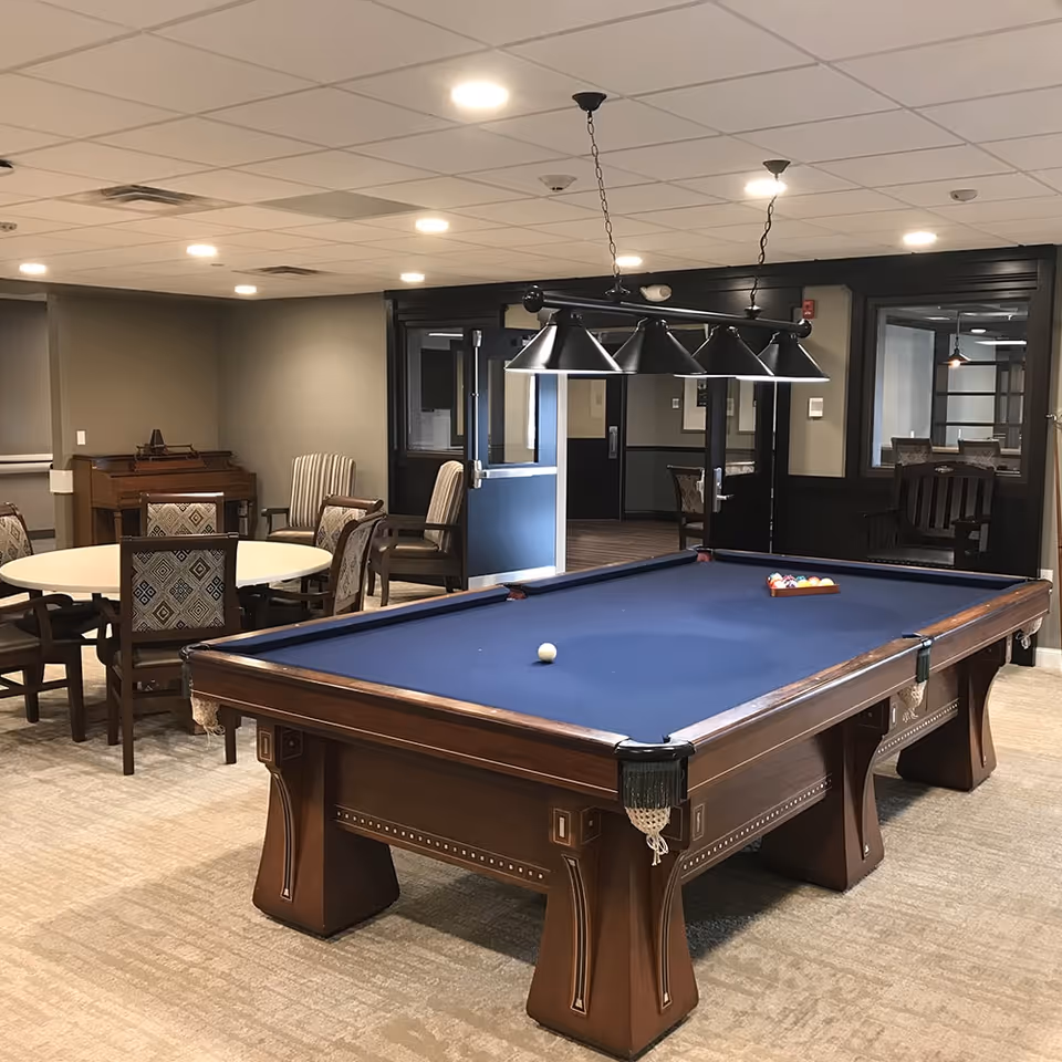 A billiards table with blue felt in a well-lit common room featuring round tables and chairs.