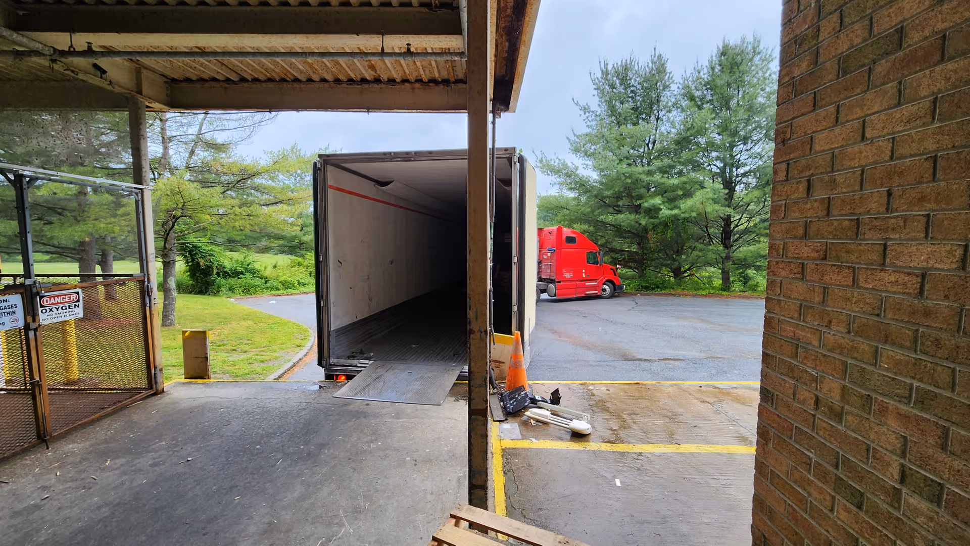 Open trailer at a loading dock beside brick wall, with a red truck in distance.